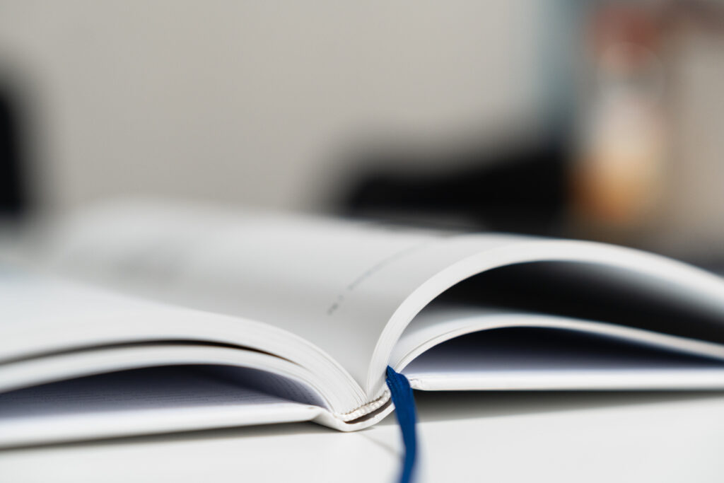 A open book with white pages and a blue ribbon bookmark, set against a blurred background, representing reading, learning, or documentation.